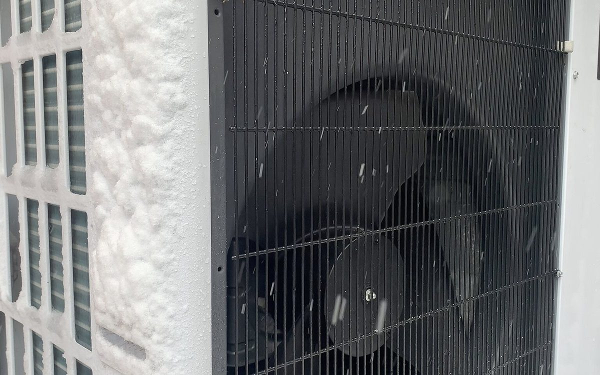 Frost and snow on the outdoor unit of an air-source heat pump beside a brick wall