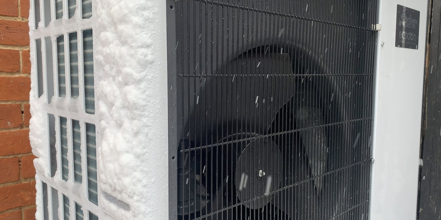 Frost and snow on the outdoor unit of an air-source heat pump beside a brick wall